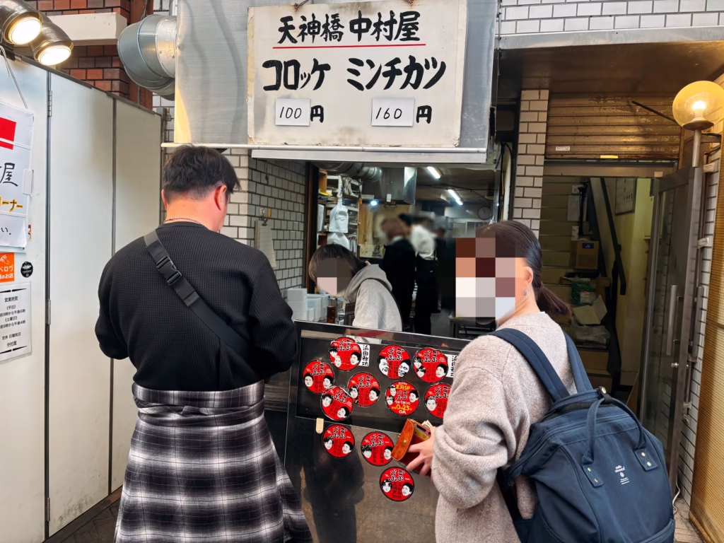 天神橋筋商店街にある天神橋中村屋のコロッケとミンチカツの表記がされたメニュー版と店内で働く人たちと購入している人たち